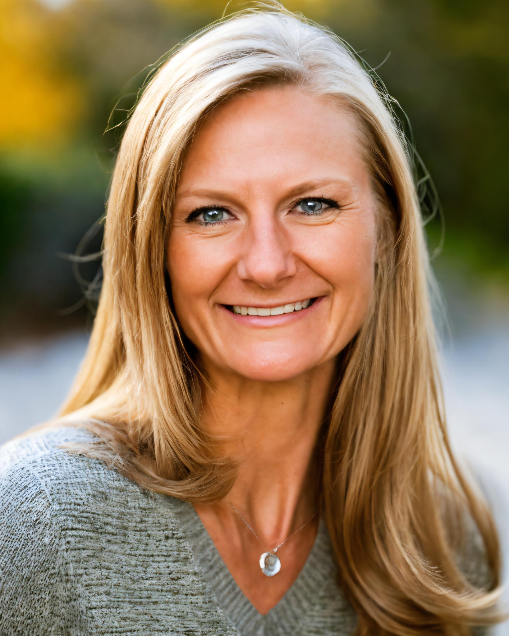 A woman with long blonde hair and blue eyes smiles at the camera, wearing a gray sweater and a pendant necklace, with a blurred outdoor background.