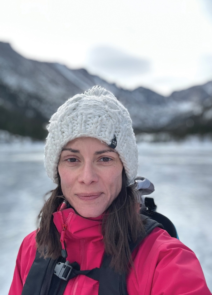 A woman in a white knit hat and red jacket stands outdoors with snowy mountains and a frozen lake in the background.