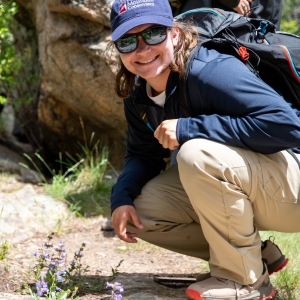 A person wearing sunglasses and a cap crouches on a rocky path outdoors, smiling at small purple wildflowers growing near the trail.