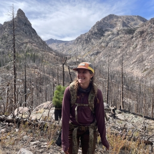 Woman wearing a hat and hiking gear stands smiling among burned trees in a mountainous landscape under a partly cloudy sky.