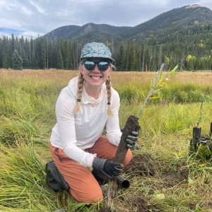 A person wearing a hat, sunglasses, and gloves kneels in a grassy field, holding a young tree seedling with exposed roots, with mountains and trees in the background.