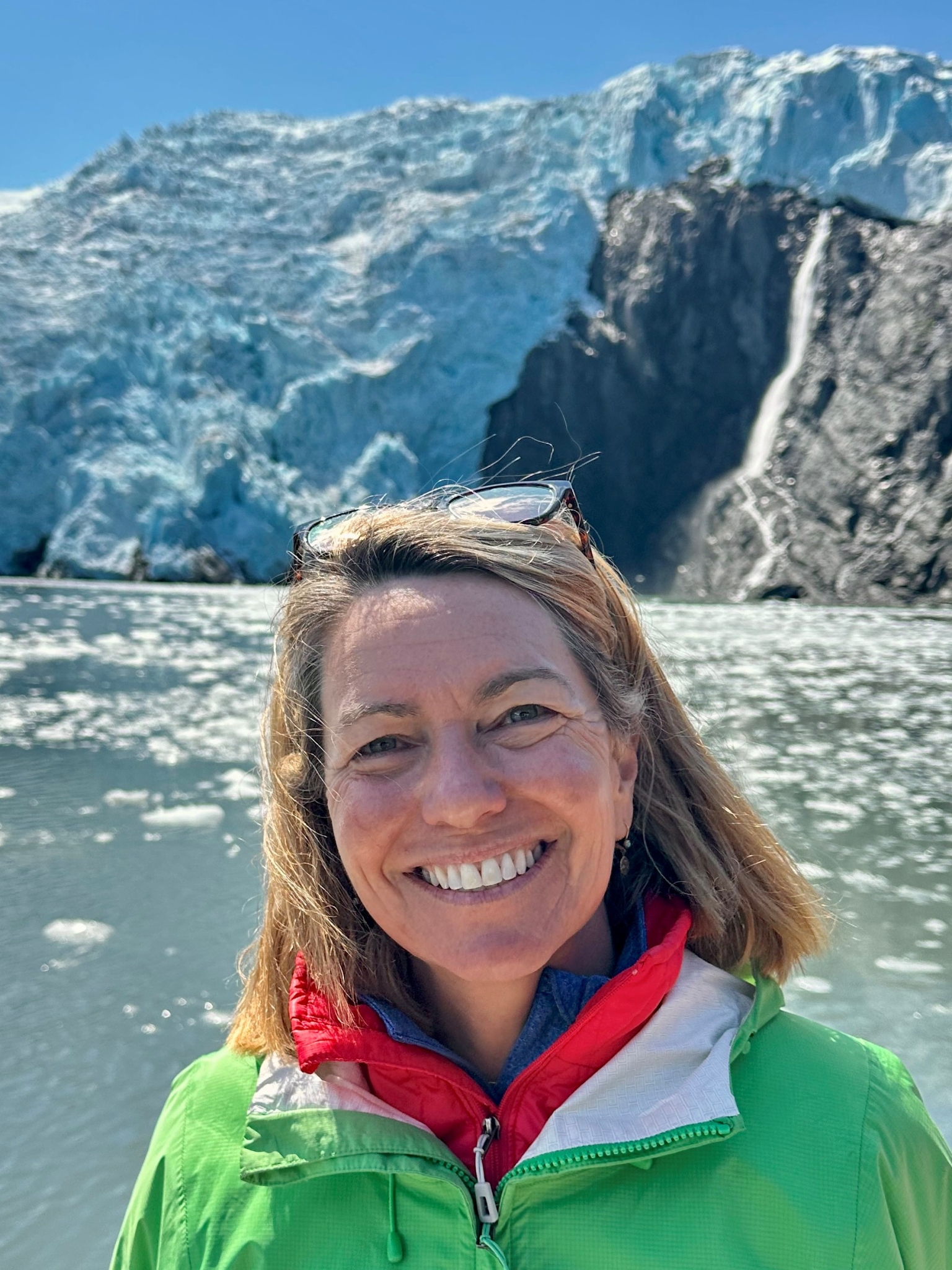 A woman wearing a green jacket smiles in front of a glacier and icy water on a sunny day.