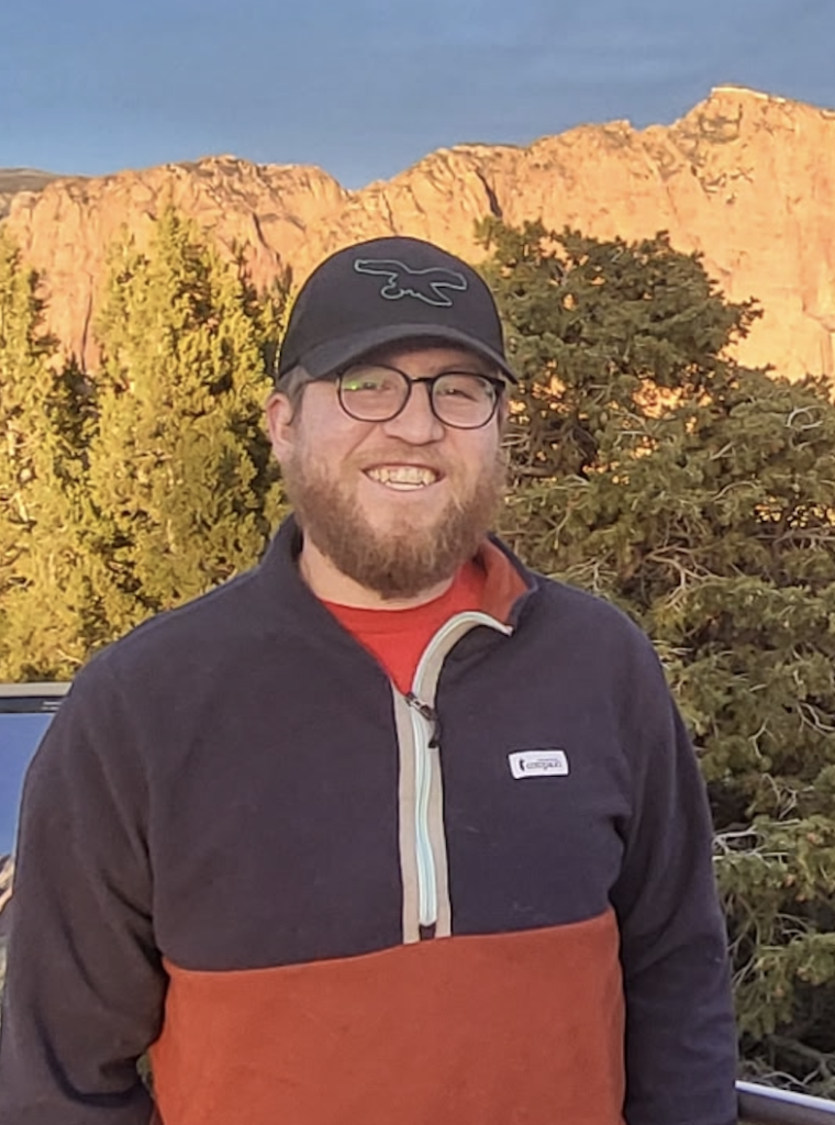 A man in glasses and a baseball cap smiles outdoors with sunlit cliffs, trees, and a parked car in the background.