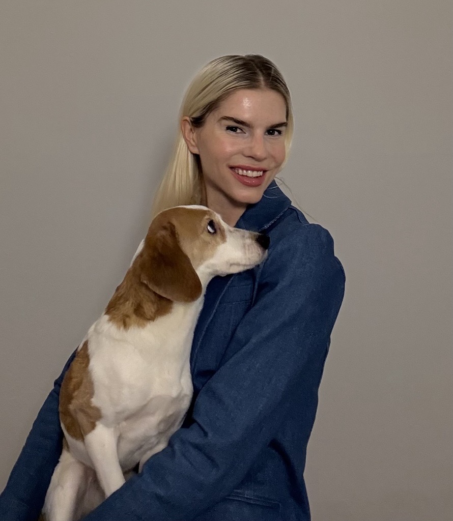 A person with long blonde hair wearing a blue top holds a brown and white dog, standing against a plain, light-colored background.