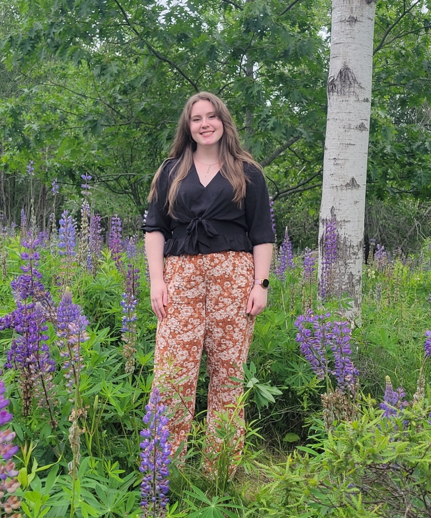 A woman stands in a field of purple wildflowers and greenery, wearing a black top and patterned orange pants, with trees in the background.
