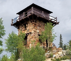 A stone fire lookout tower with a wooden balcony stands on a rocky hill, surrounded by trees under a cloudy sky.