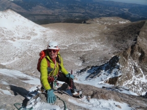 A climber wearing a helmet and winter gear ascends a snowy, rocky mountain slope with a vast landscape visible in the background.