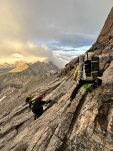 Three climbers with large backpacks and safety ropes traverse a steep, rugged granite mountain face at high altitude under a cloudy sky.