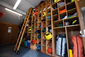 Wooden shelves with various outdoor gear, including bags, helmets, and equipment, organized in cubbies; a wooden ladder leans against the shelves in a storage room.