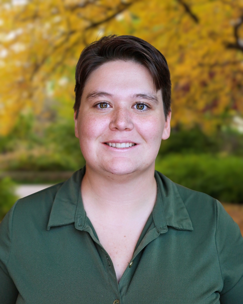 A person with short brown hair and a green collared shirt smiles outdoors with blurred autumn foliage in the background.