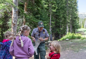 A park ranger shows an antler to three young girls in a forested area; one girl holds a magnifying glass up to her face.