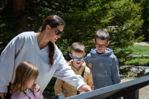 A woman and three children stand outdoors by an informational sign, looking at it together. Trees and sunlight are visible in the background.